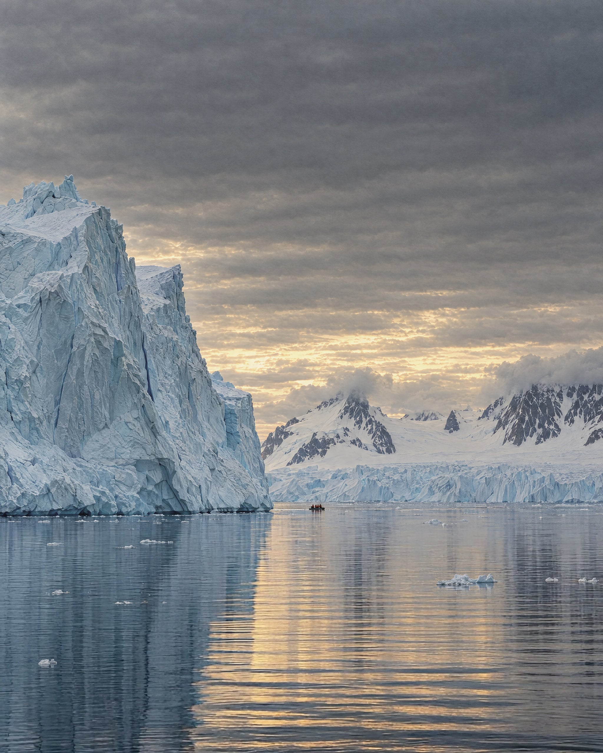 Pale blue icebergs rising from calm, dark water. A tall fractured tabular berg stands on the left, and a glacier front and snow-covered peaks with dark rock ridges sit beyond. Low grey cloud breaks to pale gold light above the horizon. Far out on the still water, a small inflatable boat carries a few passengers in red jackets.