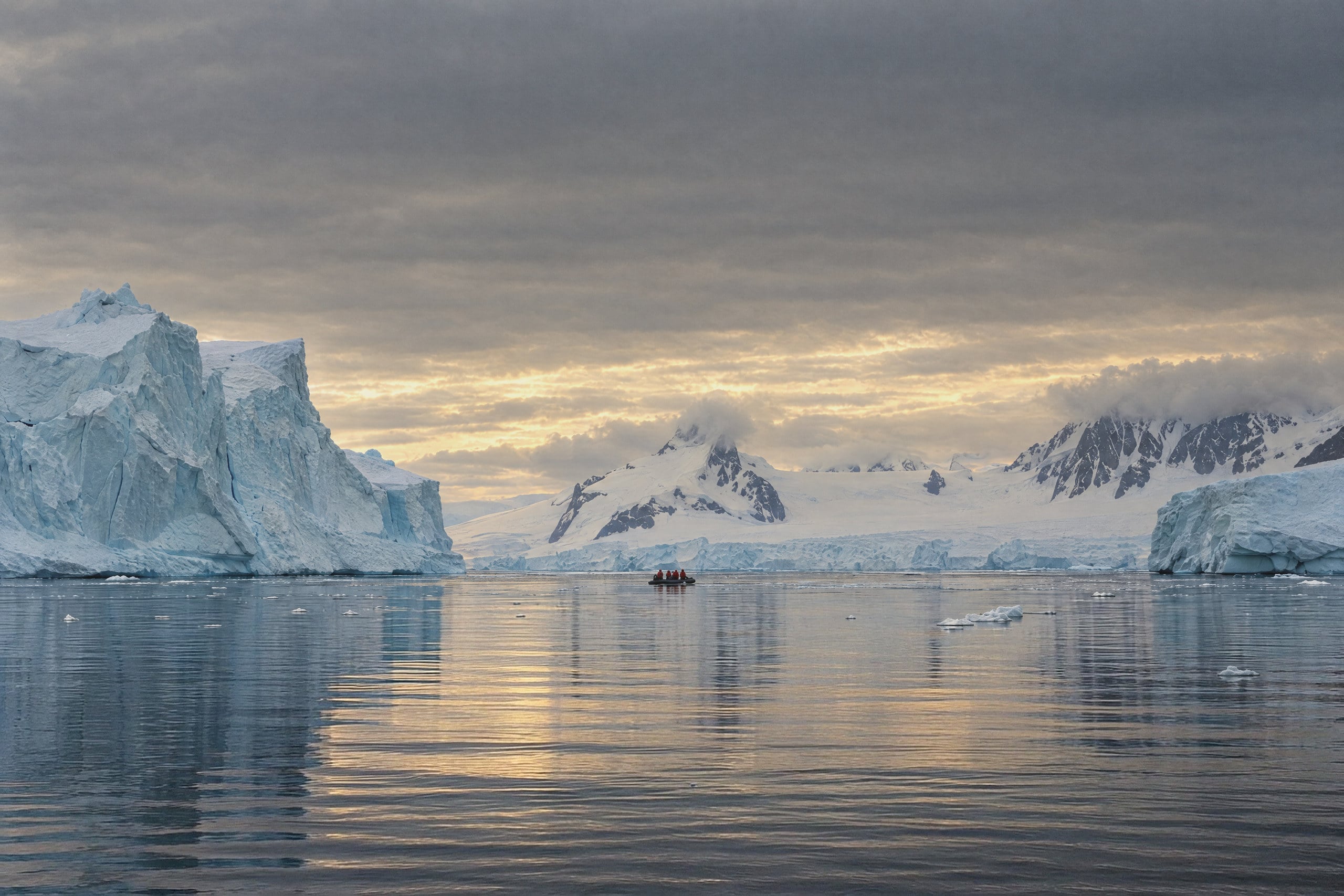 Pale blue icebergs rising from calm, dark water. A tall fractured tabular berg stands on the left, and a glacier front and snow-covered peaks with dark rock ridges sit beyond. Low grey cloud breaks to pale gold light above the horizon. Far out on the still water, a small inflatable boat carries a few passengers in red jackets.