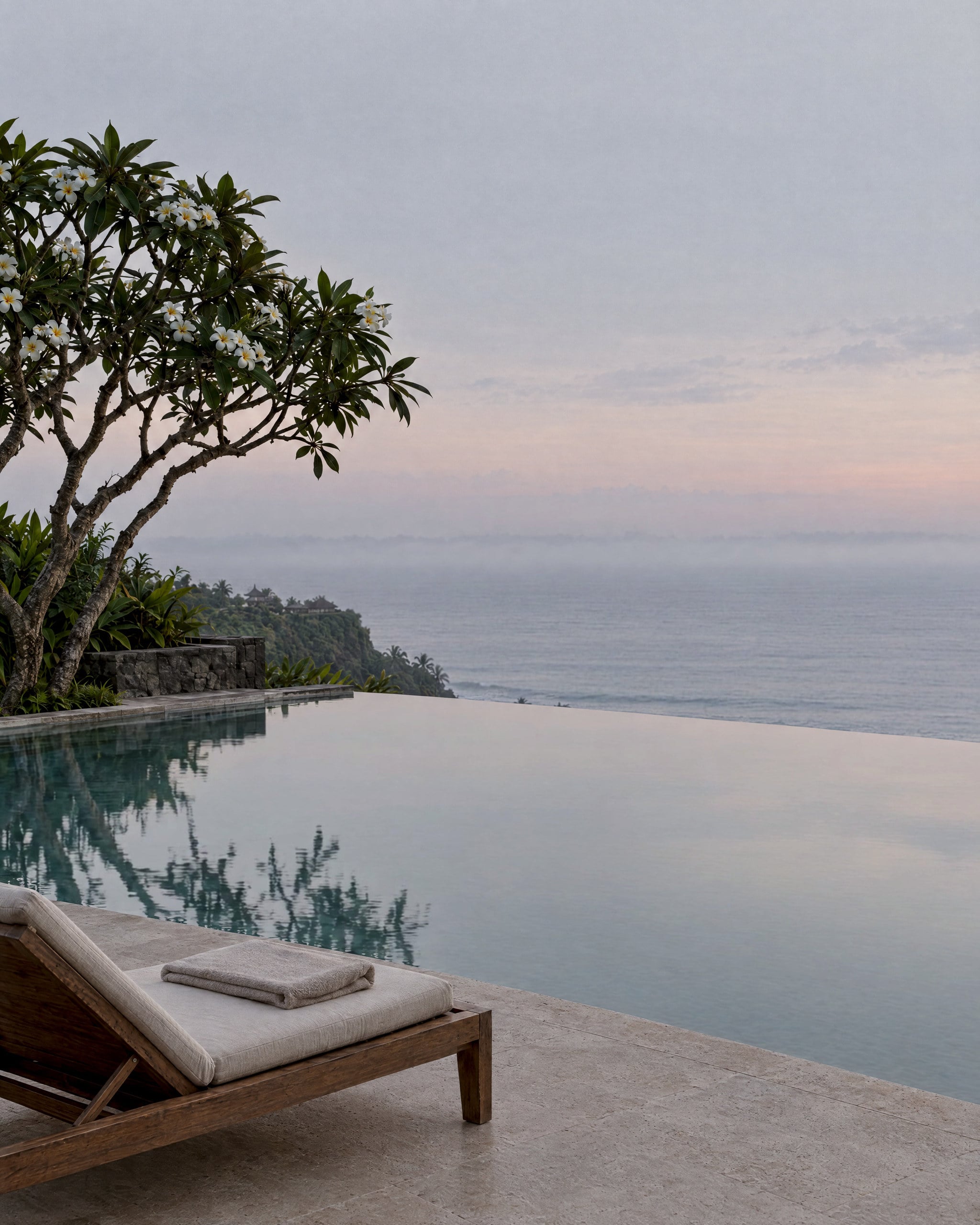 An infinity pool at first light, its still pale water mirroring a pearl-grey sky with a faint blush, beside a teak lounger with a folded linen towel on a stone deck, beneath a flowering frangipani tree, with a green headland and a thin band of mist lying over the sea.