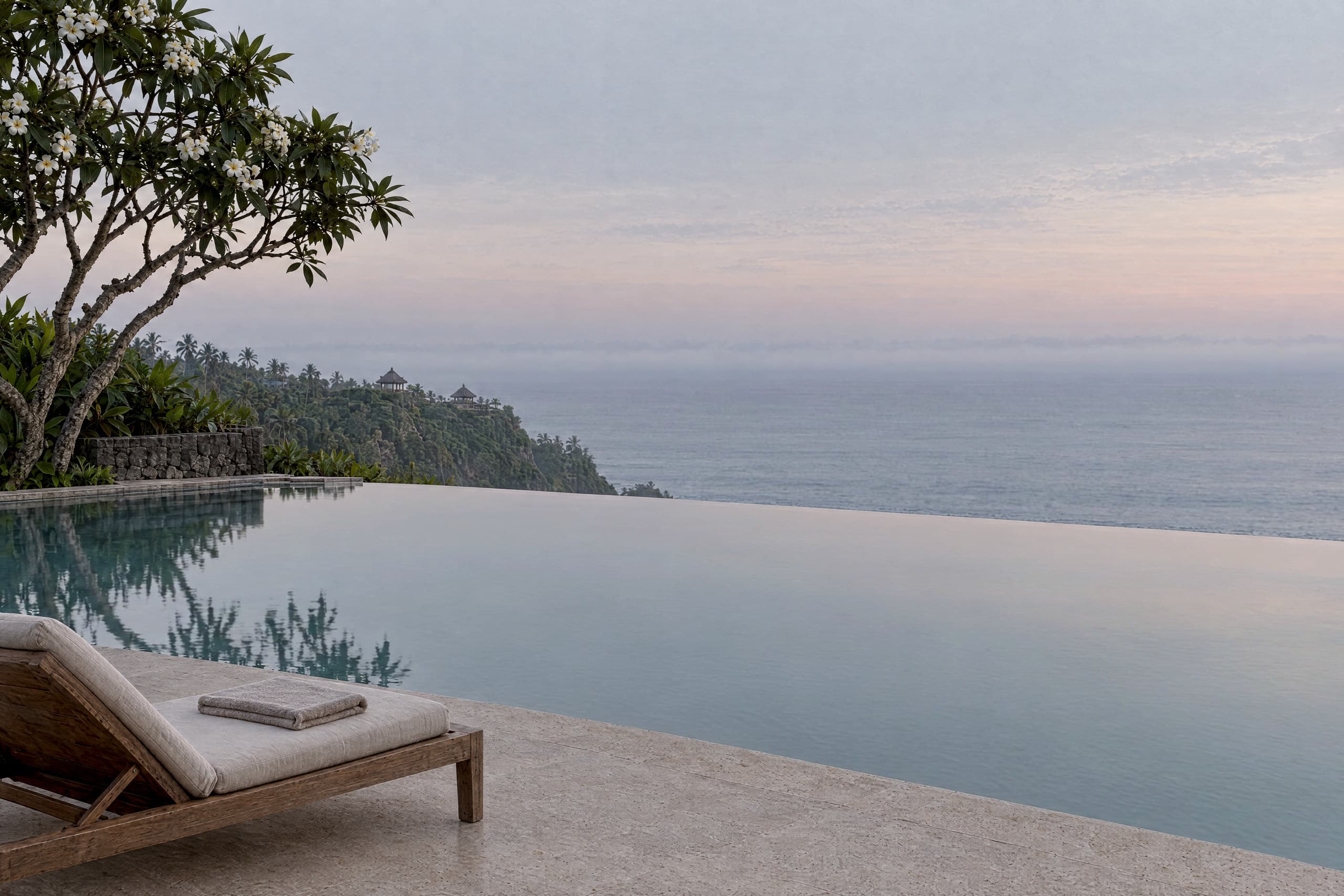 An infinity pool at first light, its still pale water mirroring a pearl-grey sky with a faint blush, beside a teak lounger with a folded linen towel on a stone deck, beneath a flowering frangipani tree, with a green headland and a thin band of mist lying over the sea.