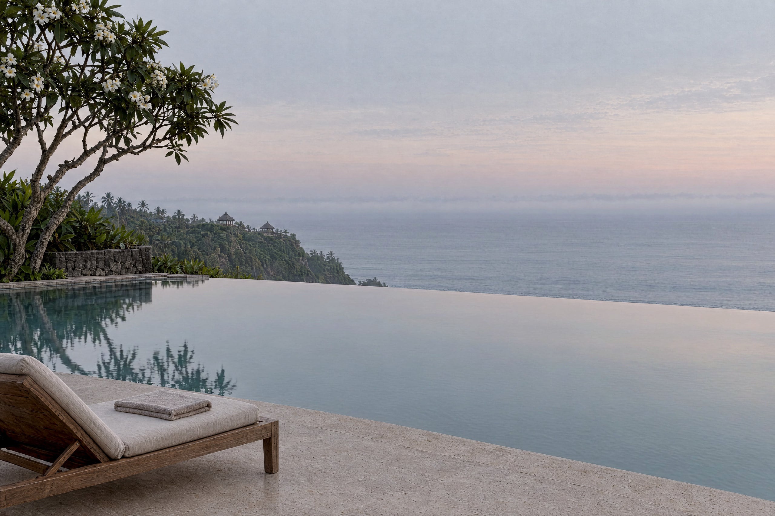 An infinity pool at first light, its still pale water mirroring a pearl-grey sky with a faint blush, beside a teak lounger with a folded linen towel on a stone deck, beneath a flowering frangipani tree, with a green headland and a thin band of mist lying over the sea.