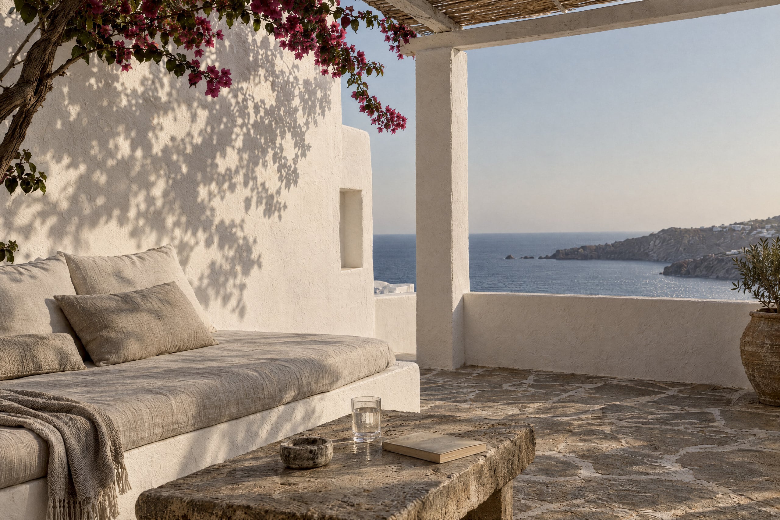 A shaded whitewashed terrace with a built-in linen daybed, a stone table with a glass of water and a book, bougainvillea casting leaf shadows on the wall, and the sea and a headland beyond.