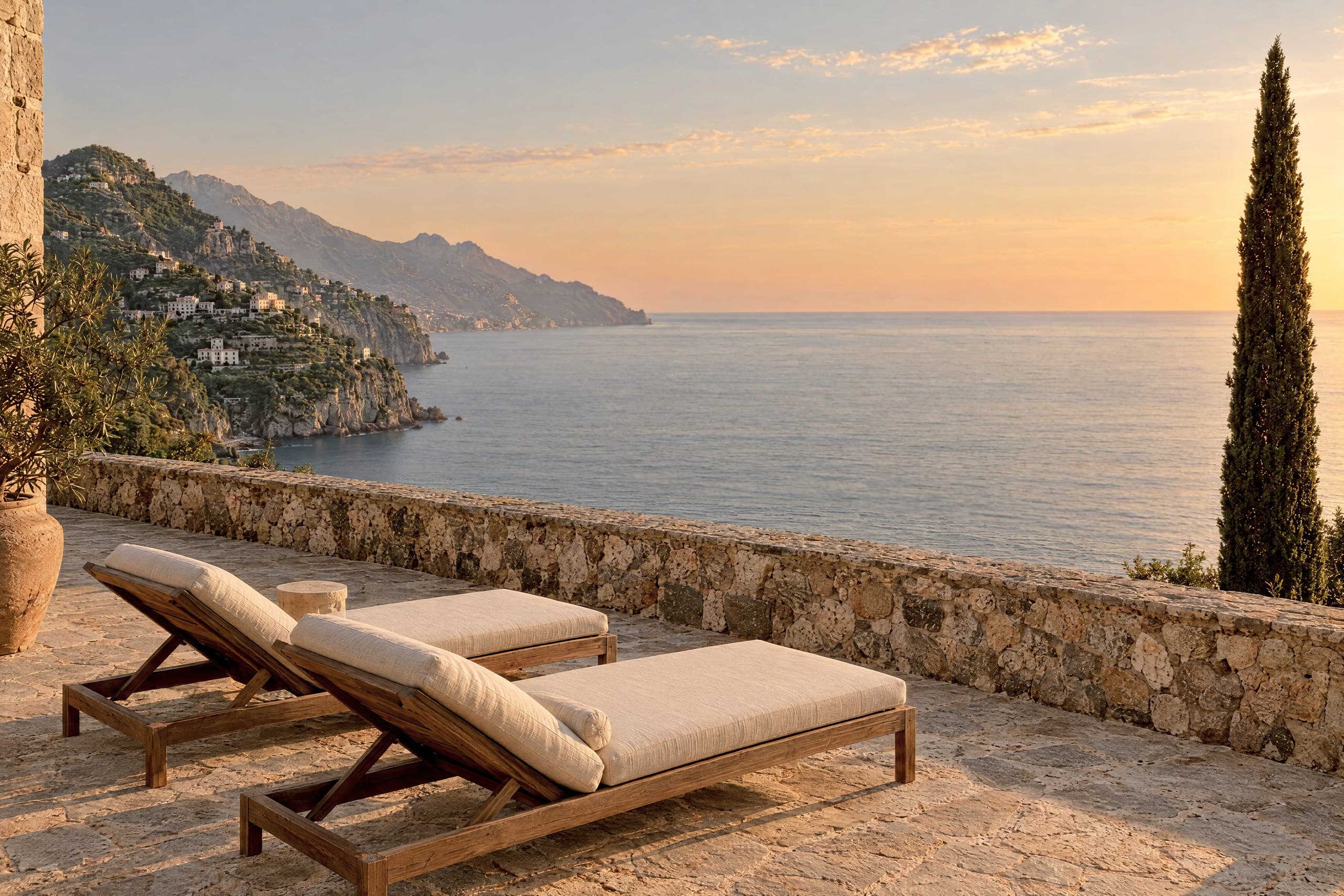 A stone terrace in low evening sun with two cushioned wooden loungers in front of a low stone wall. A tall cypress stands at the right edge. Beyond the wall, a calm sea stretches to the horizon beneath steep cliffs and a small town of pale buildings.